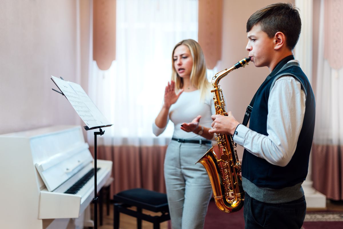 A teen playing the saxophone during a private saxophone lesson with a teacher clapping to a beat in the background